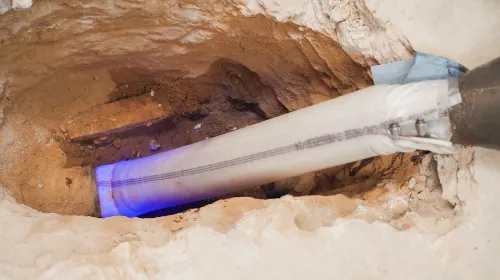 Close-up view of a trench with an exposed pipe undergoing a repair or lining process, illuminated by a blue curing light.